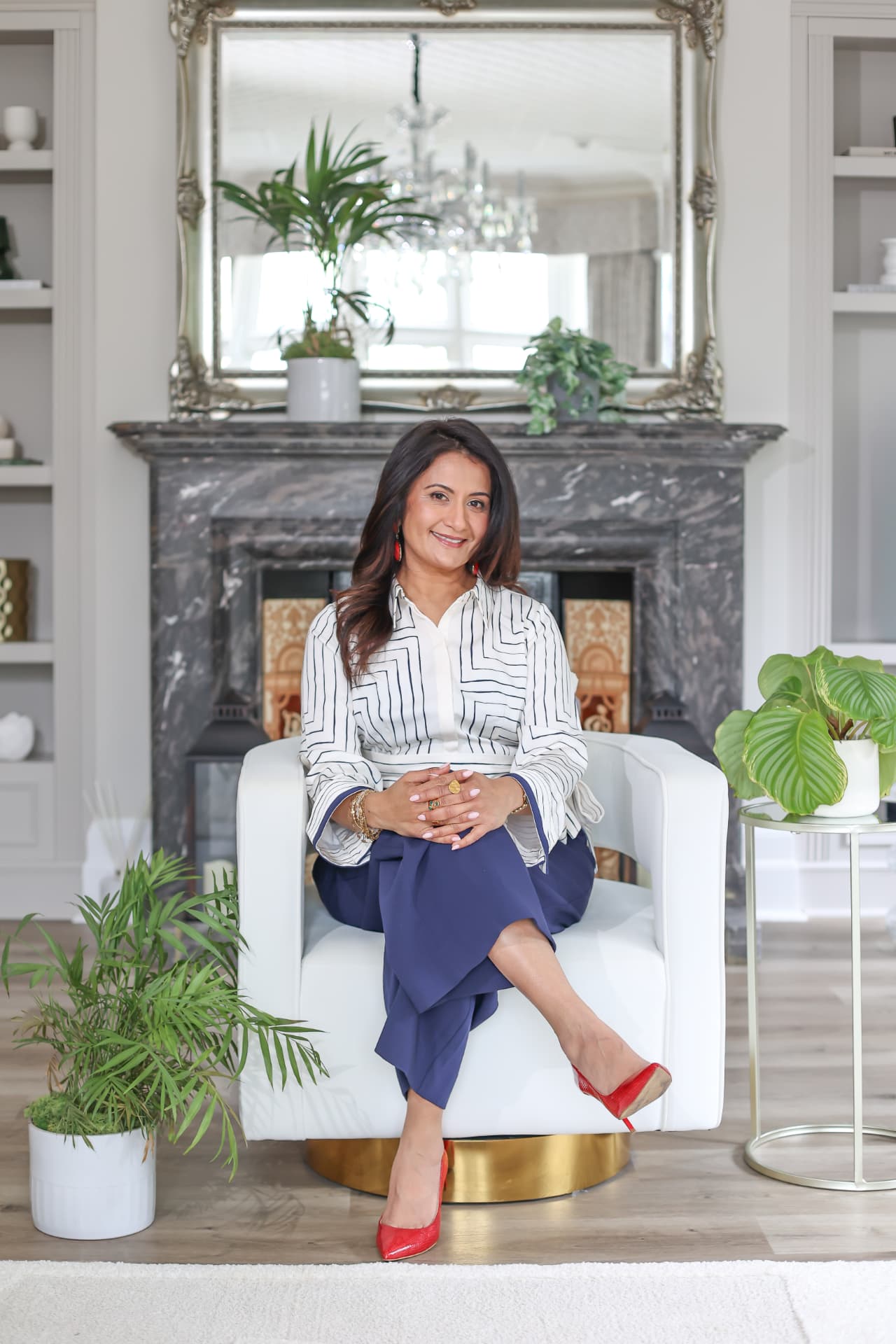 Kajal seated in a pale armchair in front of a marble fireplace, hands resting together, looking towards the camera